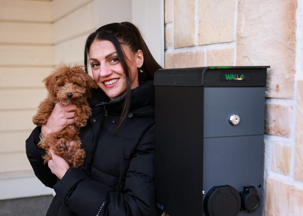 Nooralotta ja maltipoo-pentu Ato poseeraavat kotipihan sähköauton latauspisteellä. Etualalla kahden sähköauton latausasema TuplaWalle.